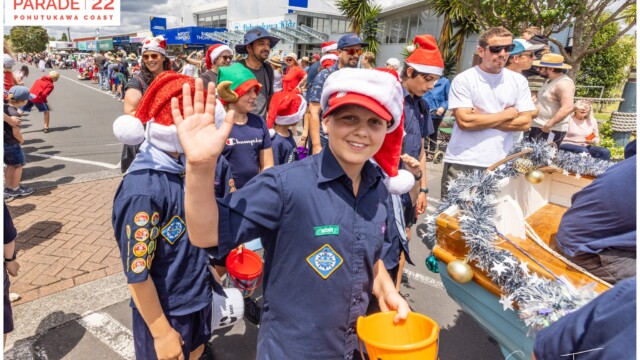 Image © Claude Thérond – used with permission Scout waving at camera during Pohutukawa Coast Santa Parade 2022, wearing Santa hat and holding collection bucket